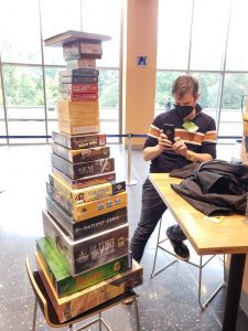 A tall stack of board games, with the author in the background photographing them.