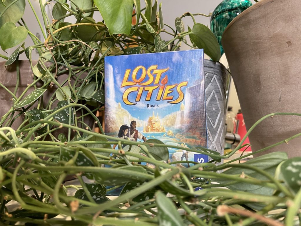 The box for Lost Cities: Rivals tastefully arranged in some shubbery.