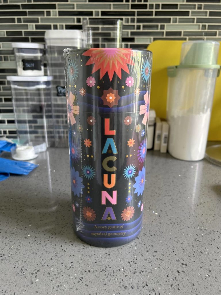The Lacuna packaging, a cardboard tube with a black background and vibrant, round shapes all around. The title of the game is arranged in a vertical column.