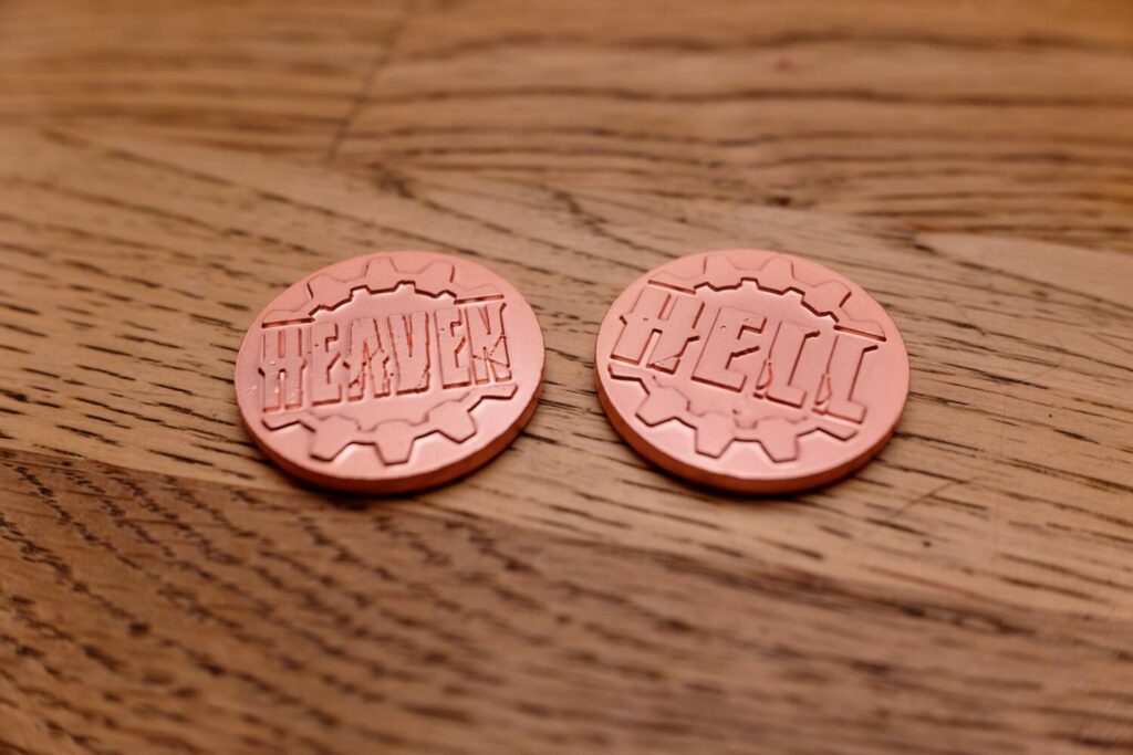 The health markers for each player are metal coins with the words "Heaven" and "Hell" engraved into them.