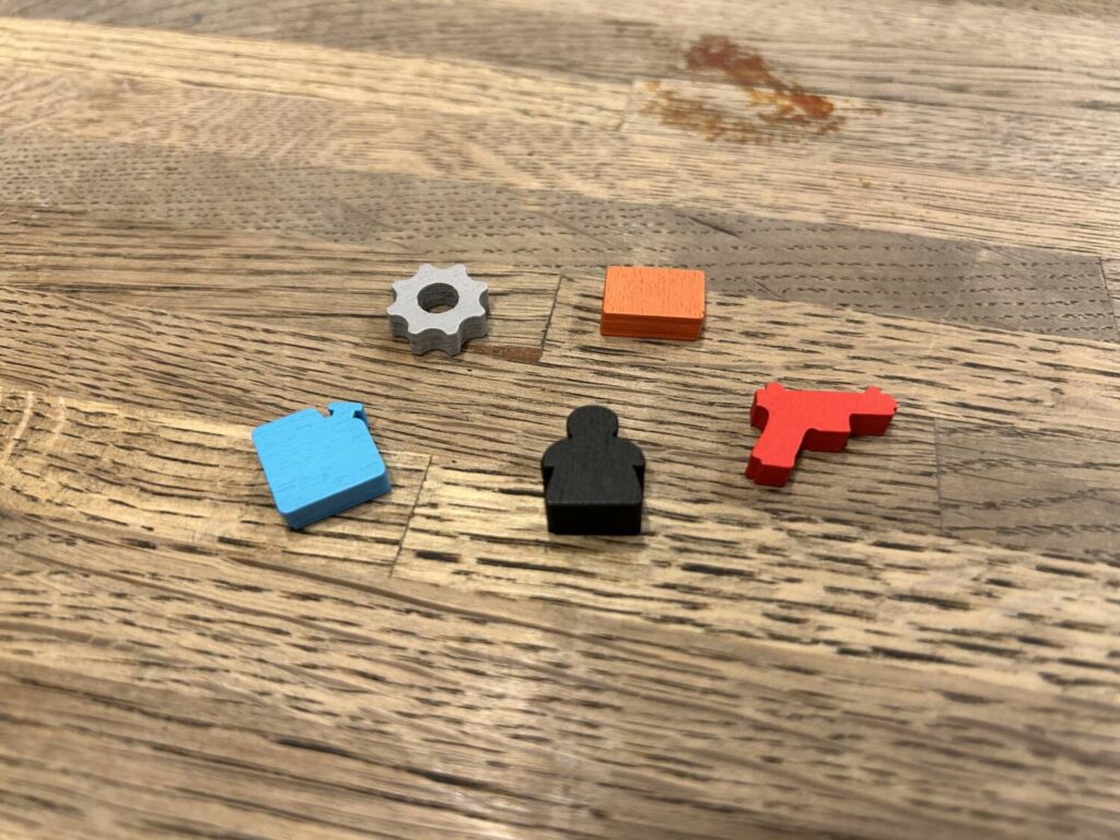 Five wooden resource markers sit in a loose circle on the table. They include a grey cog, an orange brick, a red pistol, a black meeple with a somewhat atypical shape, and a blue gas canister.