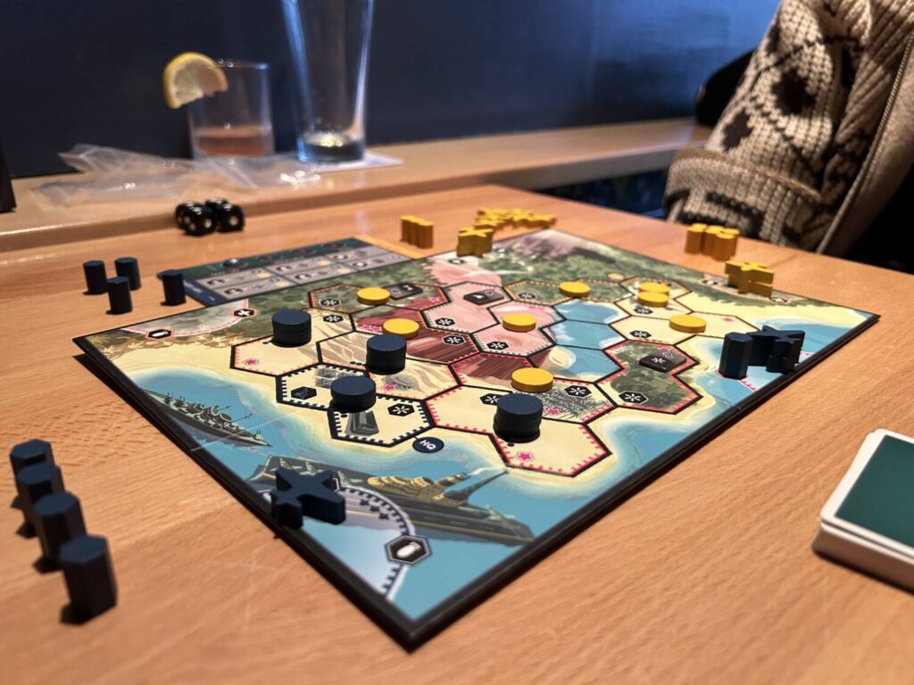 The game board is covered in hexagonal spaces. The player components are pieces of blue or yellow wood, primarily discs.