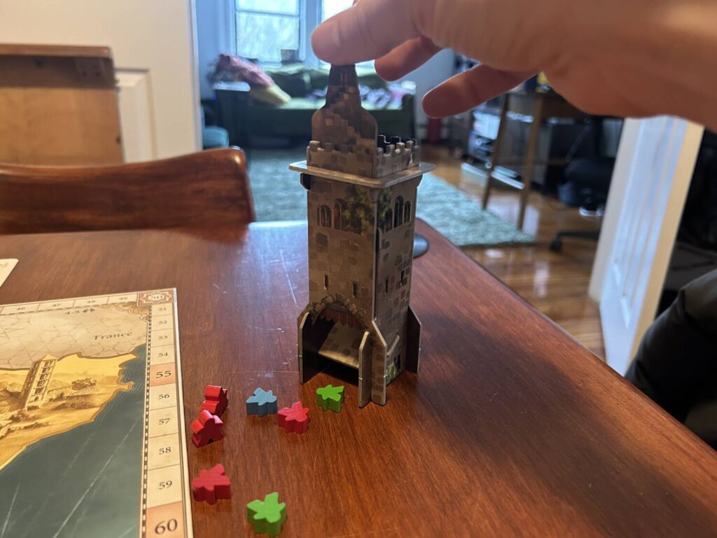 A hand reaches into frame to lift the gate on the El Grande tower. Meeples spill out onto the table.