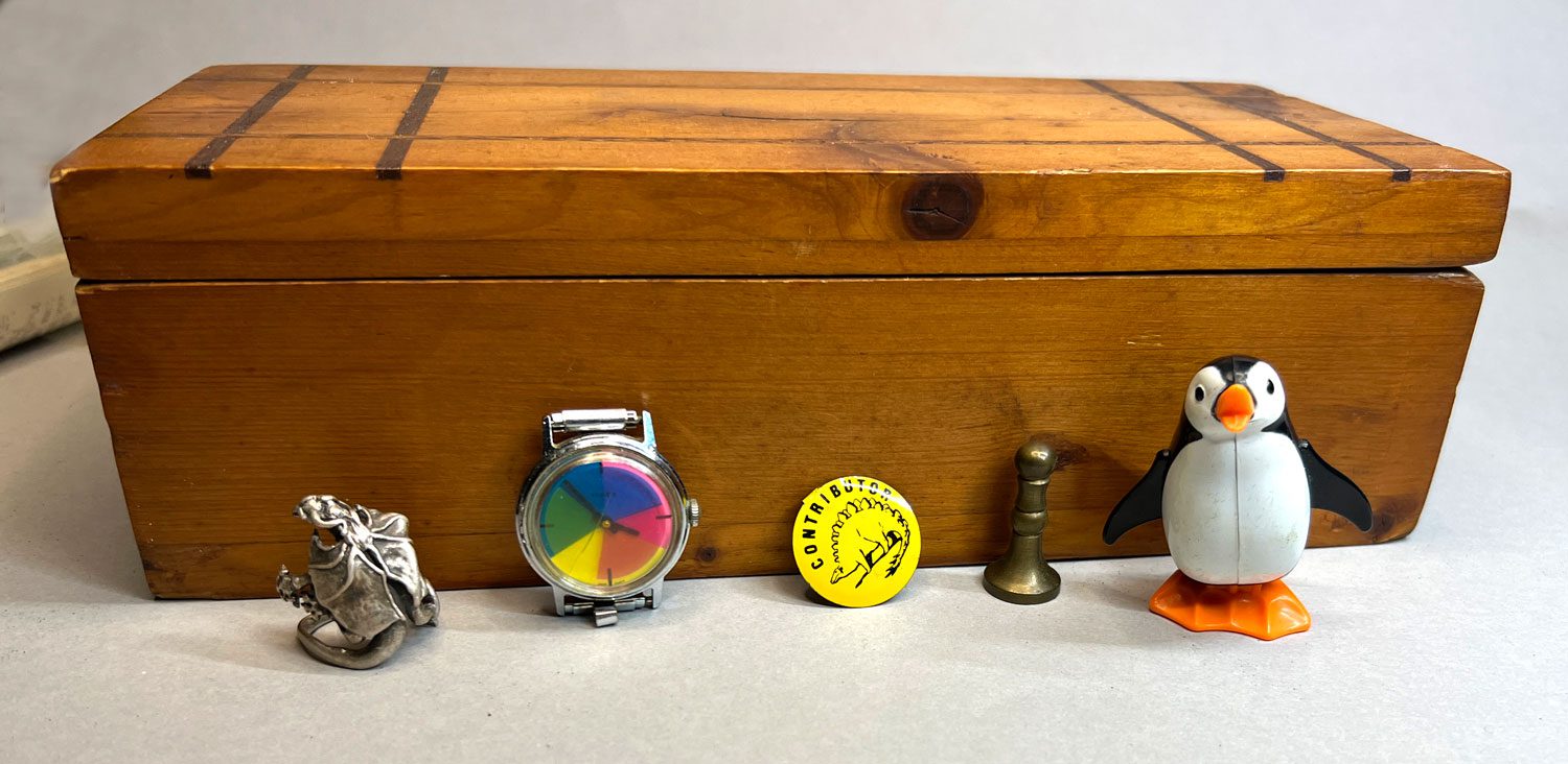 My Jr. High School box holding up my collection of player pieces: my silver dragon ring, Twinkle's watch, a Contributor's badge I got somewhere, a brass sealing wax impression pawn, and a wind-up penguin.
