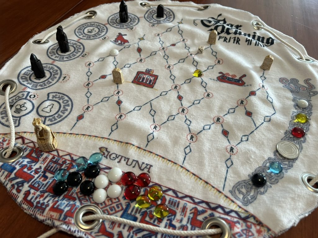 The board is a circular piece of felt, with a lattice of spaces in the middle and seven circular bowls to the left. The bottom of the board is covered with dense decorations.