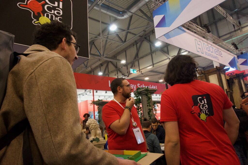 Jim speaks to two representatives at the Gigamic booth, who look over the booth personnel to figure out with whom he should speak.