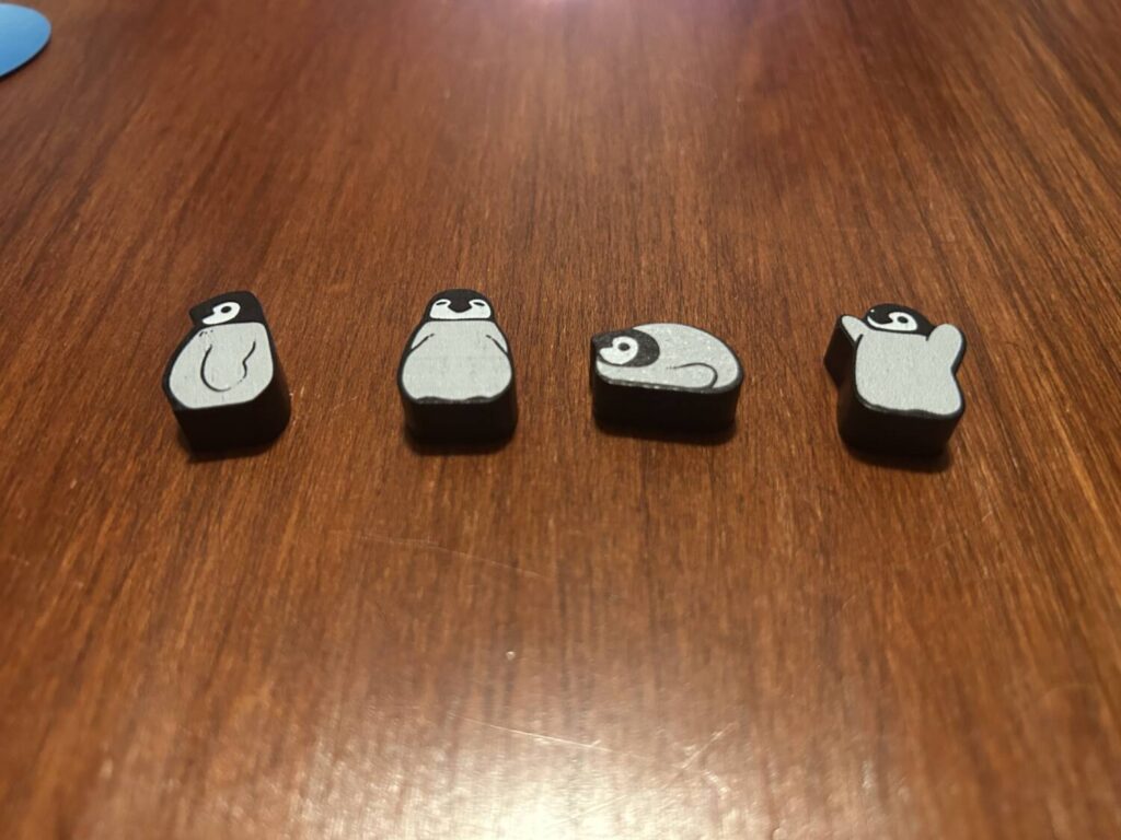 Four screen printed penguin meeples sit on the table, each in a different position. The first is viewed via its side profile, the second is facing the camera, the third has done a belly flop, and the fourth has its wings raised in ecstasy and, I assume, triumph.