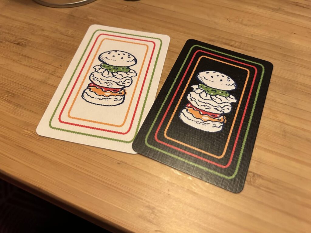 Two cards, one with a white background and one with a black background, each showing the same delicately illustrated burger, rendered with space between the ingredients, looking a bit like a cross-section illustration. The buns, patty, and lettuce are all colorless, while the relish, ketchup, and mustard are in bold colors.