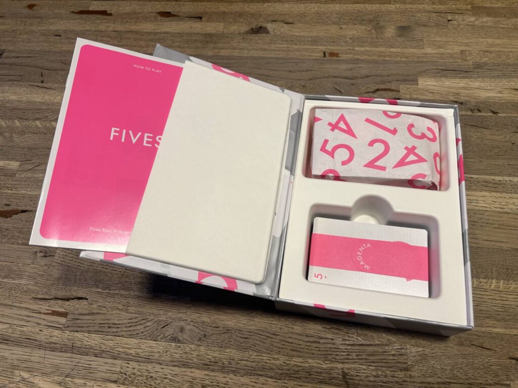 An open shot of the interior of the box for Fives. The manual is sticking out of a pocket on the interior of the lid, while the cards and tokens are in two simple trays.