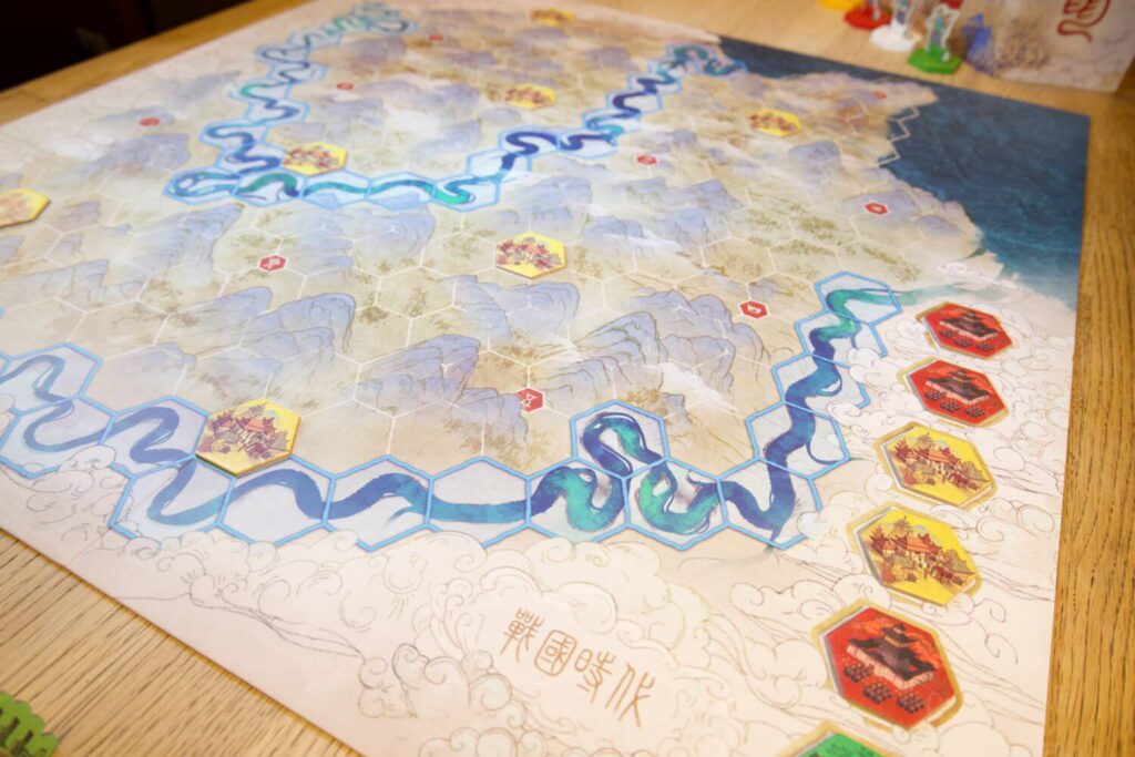 The board, illustrated to resemble an old Chinese silk screen painting, is beautiful, divided into hexagons and rich in detail, including mountain ranges and flowing rivers. The tile market sits to the right side, a column of six tiles.