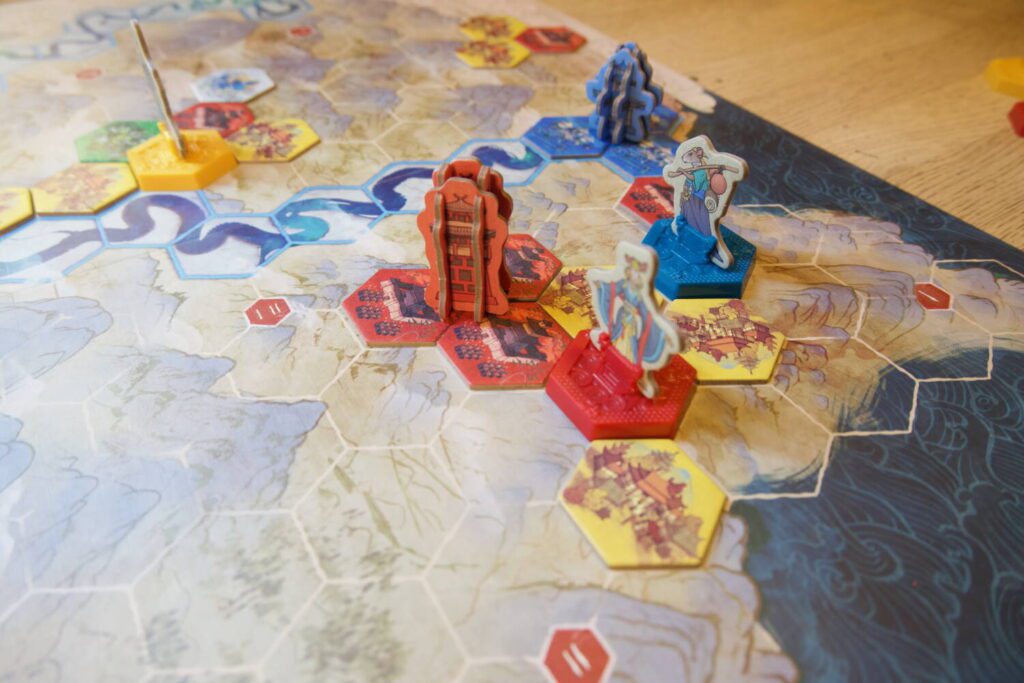 Pagodas, sturdy four-piece cardboard standees, sit at the intersection of three matching tiles. In this photo, a red pagoda sits amongst other tiles and leaders, stretching around the map. A blue pagoda sits atop blue tiles towards the bag of the image.
