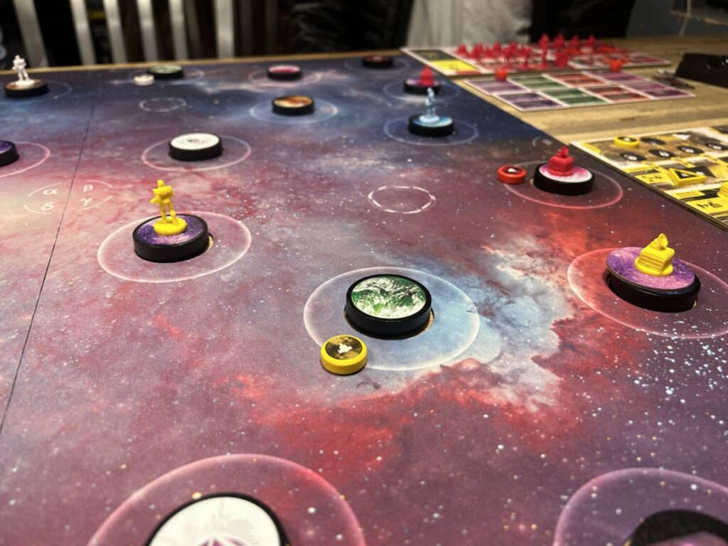 Thick wooden planet tokens sit in holes cut out of a neoprene mat, which is decorated with nebulae. There are wooden ship tokens and plastic building miniatures near and on the various planets.