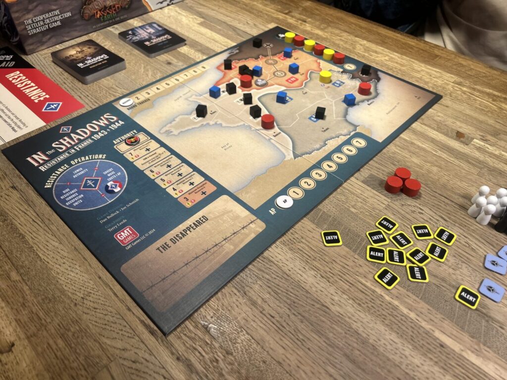 A game board on a wooden table. The board shows France, divided into multiple sections. There are numerous wooden pieces on the board. Two decks of cards sit above the board.