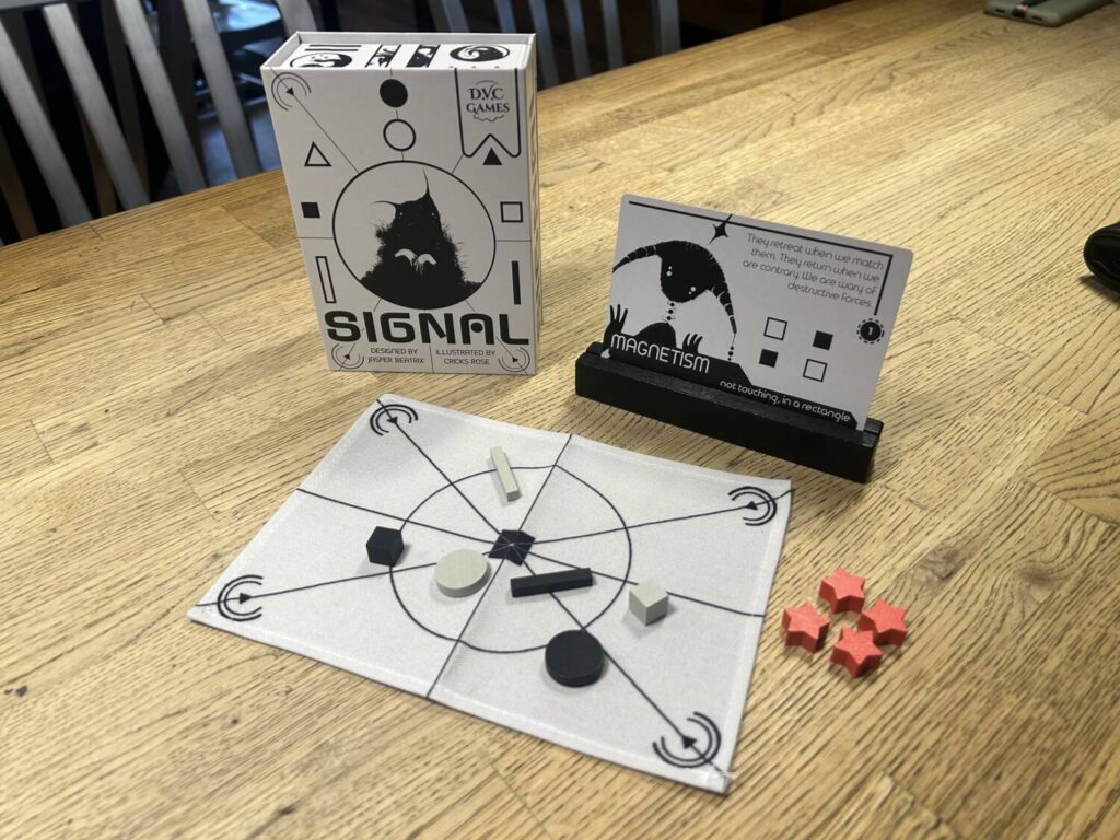 A small, white game box stands on the table next to a flashcard sitting in a wooden display tray. In front of them sits a rectangular white cloth board with wooden pieces arranged on it in a haphazard pattern. Four red wooden stars sit to the right of the board.