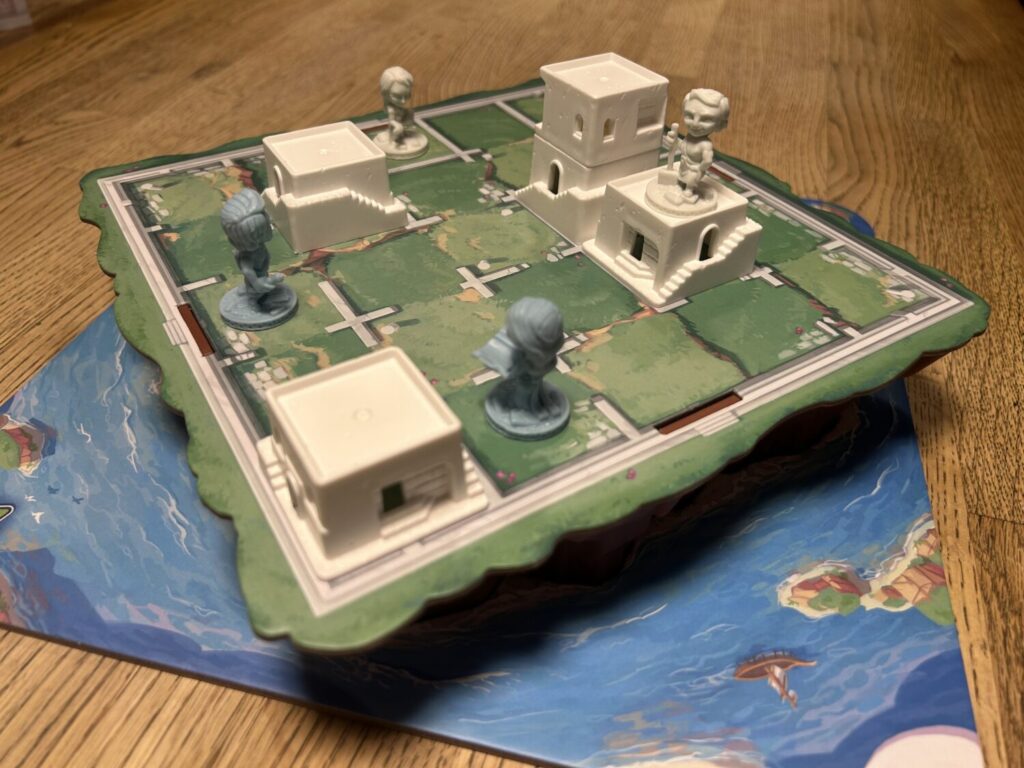 A green board atop a plastic base, molded to resemble rocky cliffs. The board is divided into a five-by-five grid, with white plastic buildings and molded sculptures of workers in various spaces.