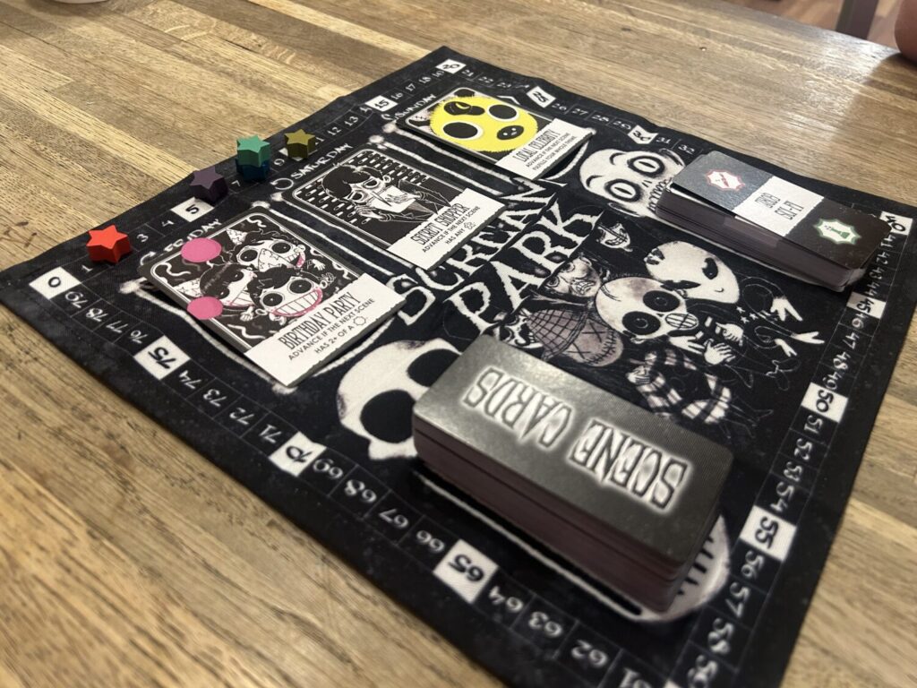 A black, square cloth board covered with white illustrations of skulls and aliens. A row of three boldly-illustrated face-up cards line the top, while two tall stacks of narrow, tall cards lay at the bottom. A score track runs along the outside.