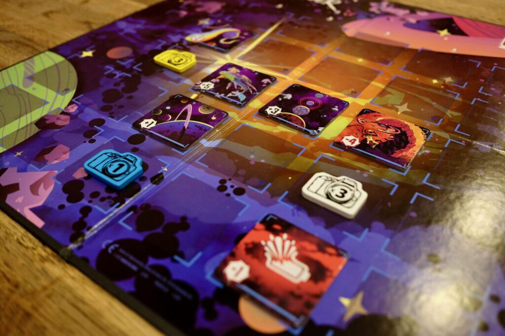 Square cardboard tiles laid out on a grid. The board shows an illustration of deep space, with purples, blacks, and yellows. There are also wooden, camera-shaped tokens on some spaces in the grid.