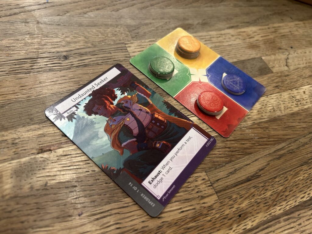Two cards on a wooden table. The ranger card is a full-body illustration of the ranger, with their ability listed at the bottom of the card. The other card, an Aspect Card, is divided into four regions, each a different color. There are circular tokens on the card.