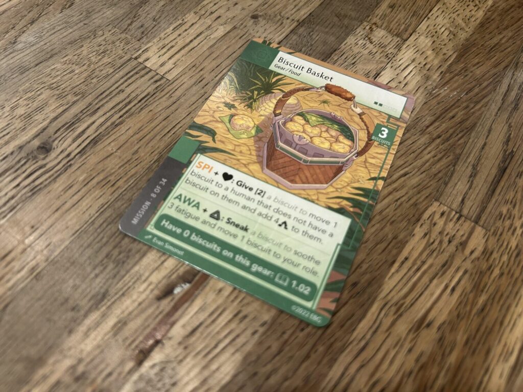 A card on a wooden table. The card is titled Biscuit Basket. It shows a drawing of a basket containing biscuits on a wooded path. A block of text occupies the bottom portion of the card.