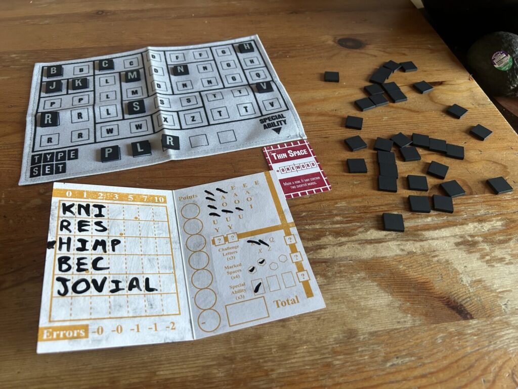 A cloth board, small black wooden tiles, a dry erase booklet with words partially written in it, and a red power card sit on a wooden table.