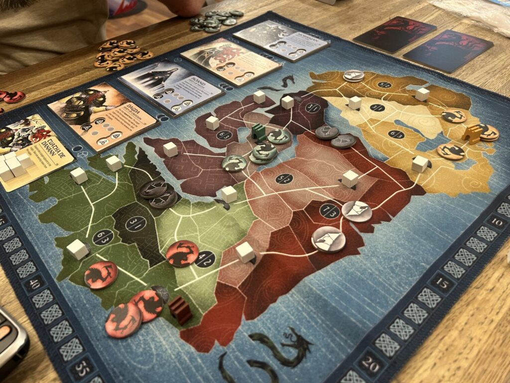 A large cloth mat with an abstracted Ireland, rendered in earthy greens, reds, purples, and yellows. Interconnected routes cut through different districts on the board, and tokens occupy various spaces. To the left, still on the board, four cardboard tiles sit in a row above a card covered in white cubes.