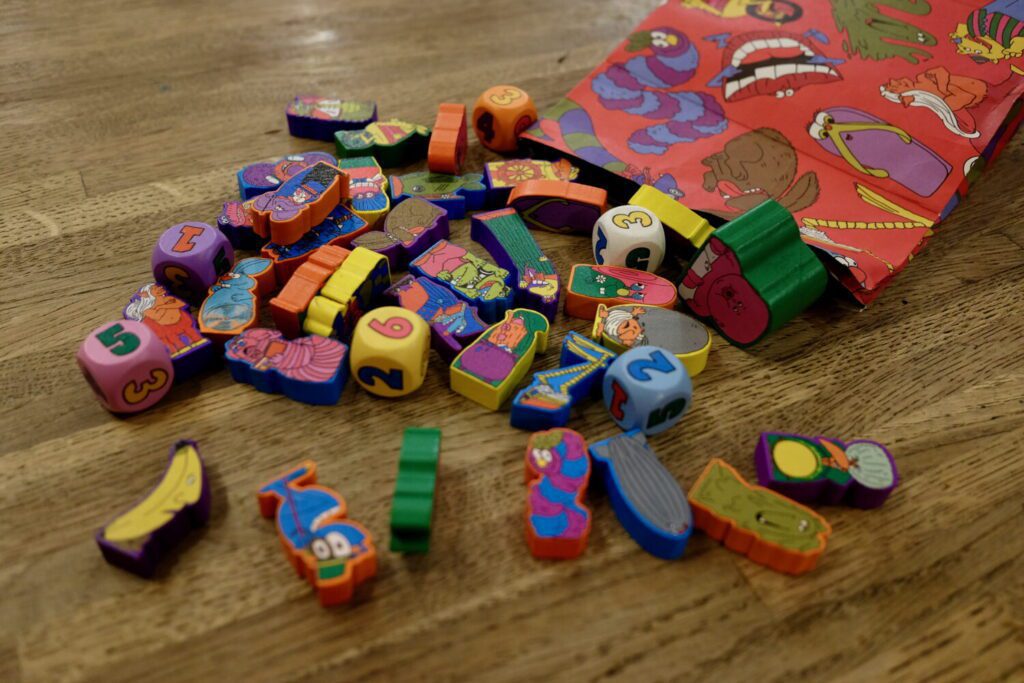 A large selection of oddly-shaped wooden pieces spread across a table, each with a full-color illustration screenprinted on both sides.
