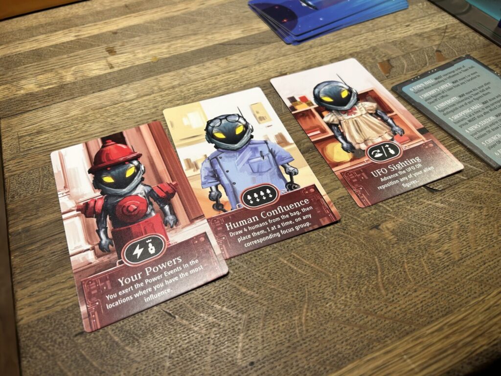 Three player cards, each decorated with an illustration of an alien in a costume on the front, sit on a table surface.