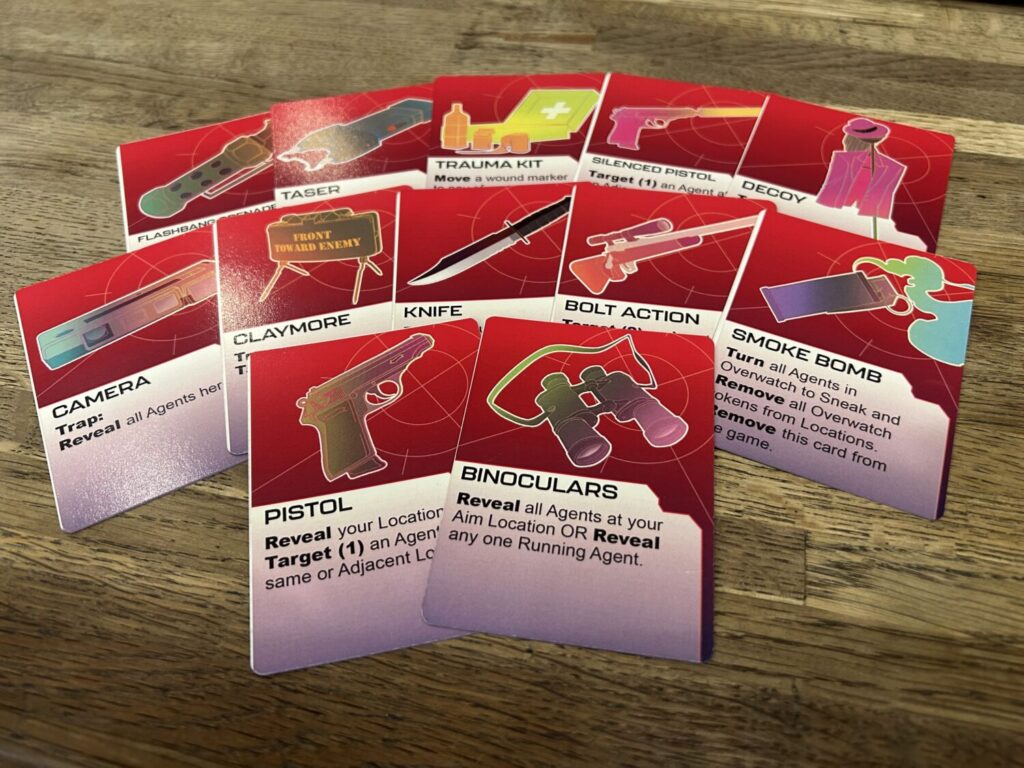 Twelve gear cards fanned out on a wooden table. Each includes an illustration of the gear against a red background and with a gray text box at the bottom of the card.