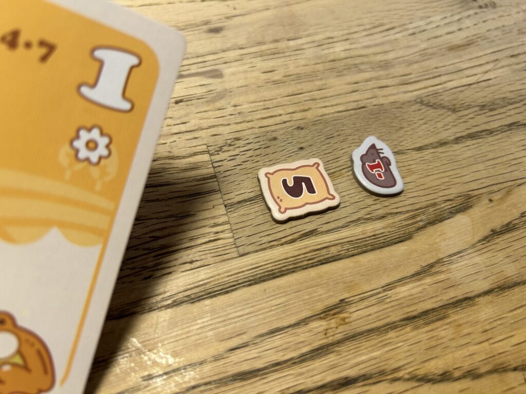 A five-point scoring token and a -1 scoring token sit on a wooden table. The 5-point token is shaped like a sack. The -1 is a mouse.