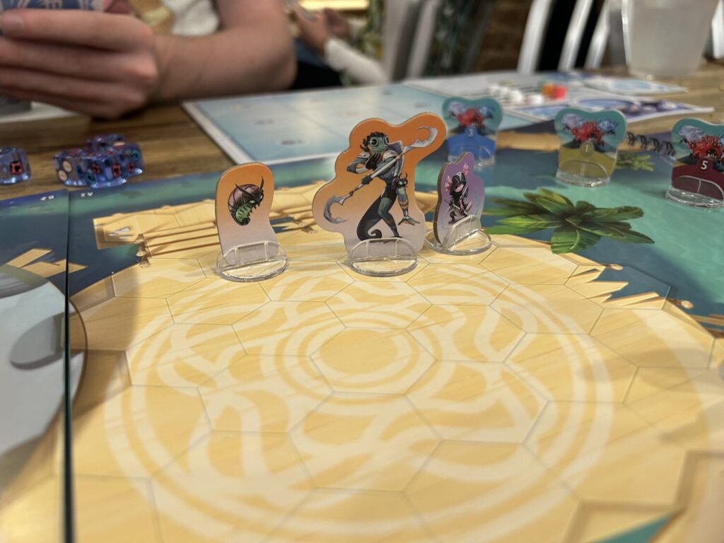 Cardboard standees on a board covered in hexagons. The illustration on the board, which is printed on one page of a spiral-bound book, shows a wooden platform out over the ocean.