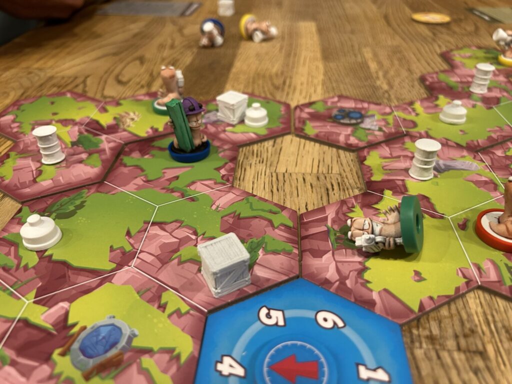 A board made up of hexagonal pieces sits on a wooden table. Plastic worm, crate, mine, and oil drum miniatures populate the board.