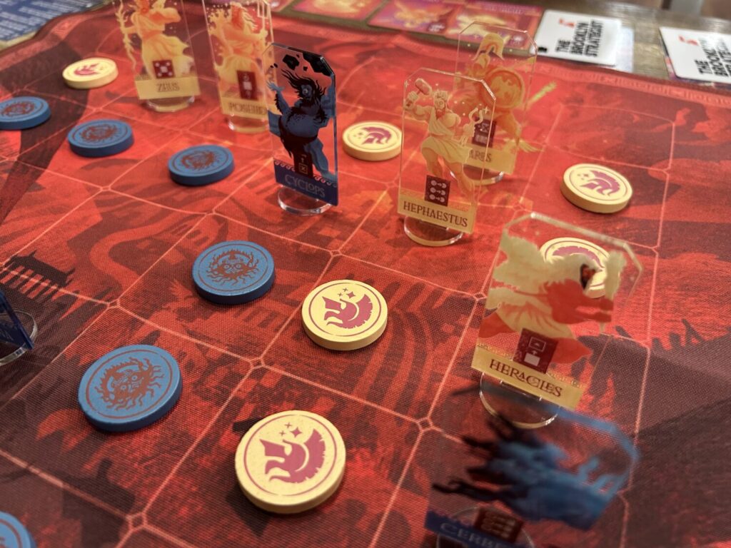 A red cloth board, divided into a 6 x 6 grid. Many of the squares are occupied by either acrylic standees or wooden tokens.