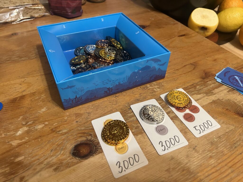 A blue box filled with coins sits on a wooden table. Three rectangular reward cards sit in front of it, each with several coins stacked on top.