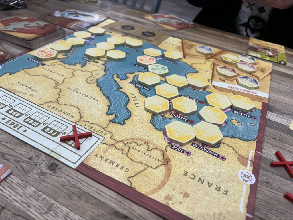 A board imprinted with a map of the Mediterranean, with many wooden hexes going across the Mediterranean sea from Africa to Europe. Most of the tiles are yellow, but a few have been flipped over to reveal the red or blue icons on the other side.