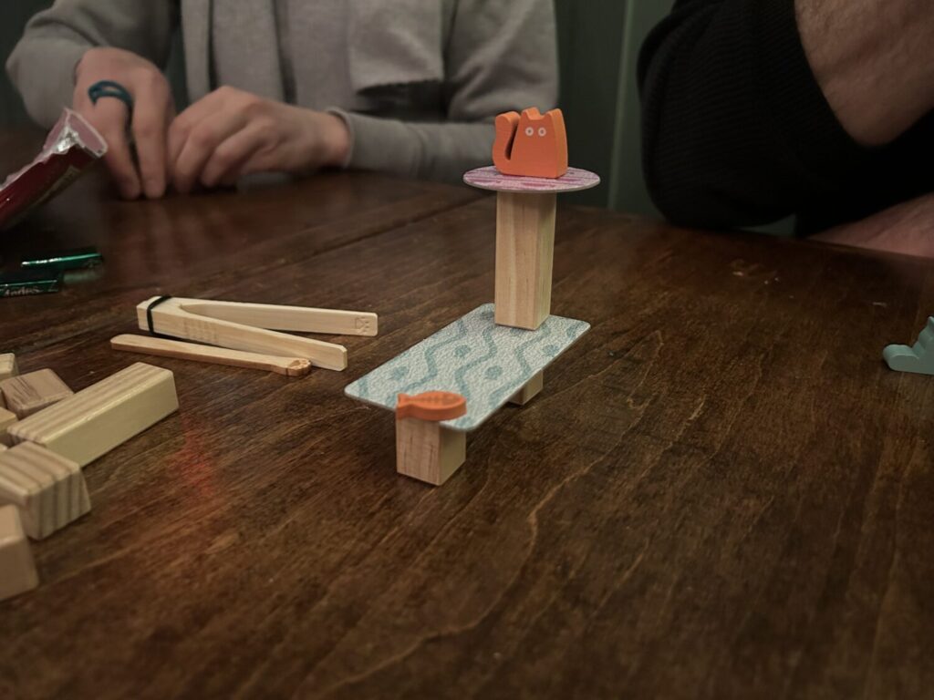 An orange wooden cat sits on top of a tall, narrow column above a wider rectangular platform, upon which sits a wooden fish.