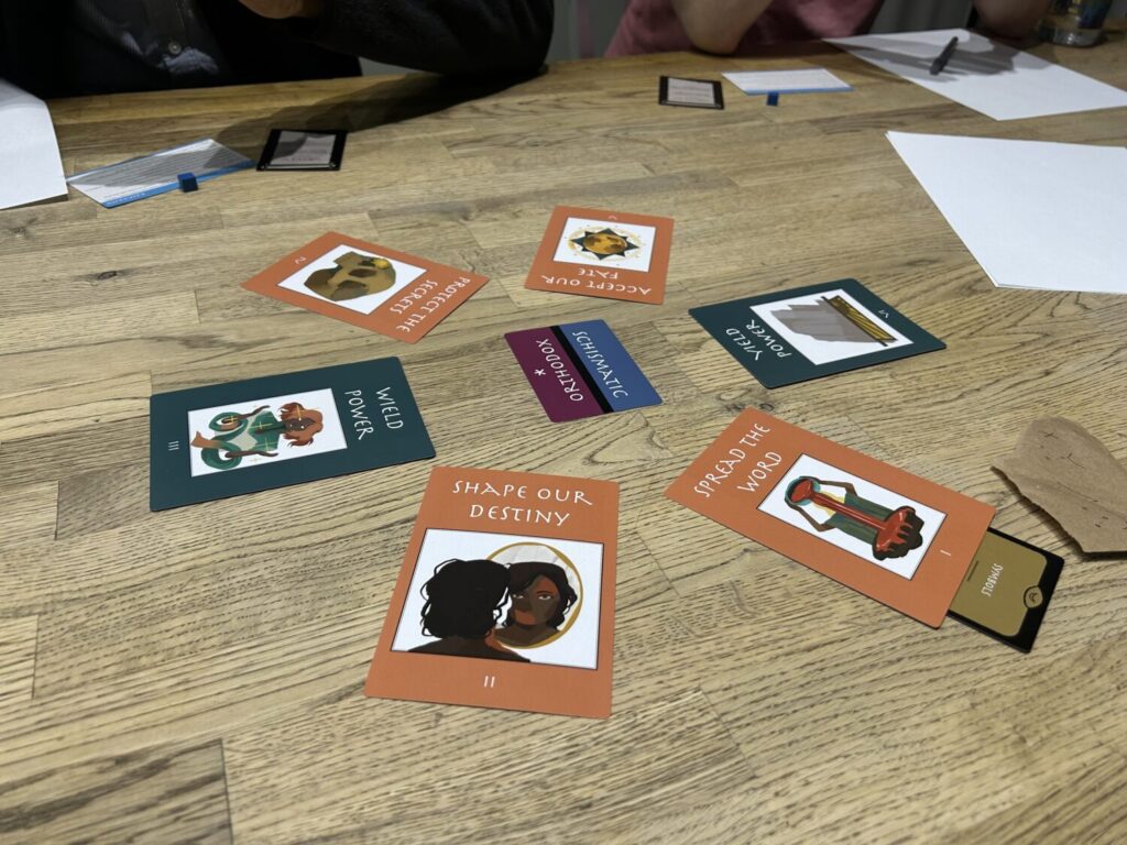 Six cards sit in a circle in the center of a table. Each has an illustration and some text at the top. The card in the middle of the circle has a line through the middle, separating the words Orthodox and Schismatic.