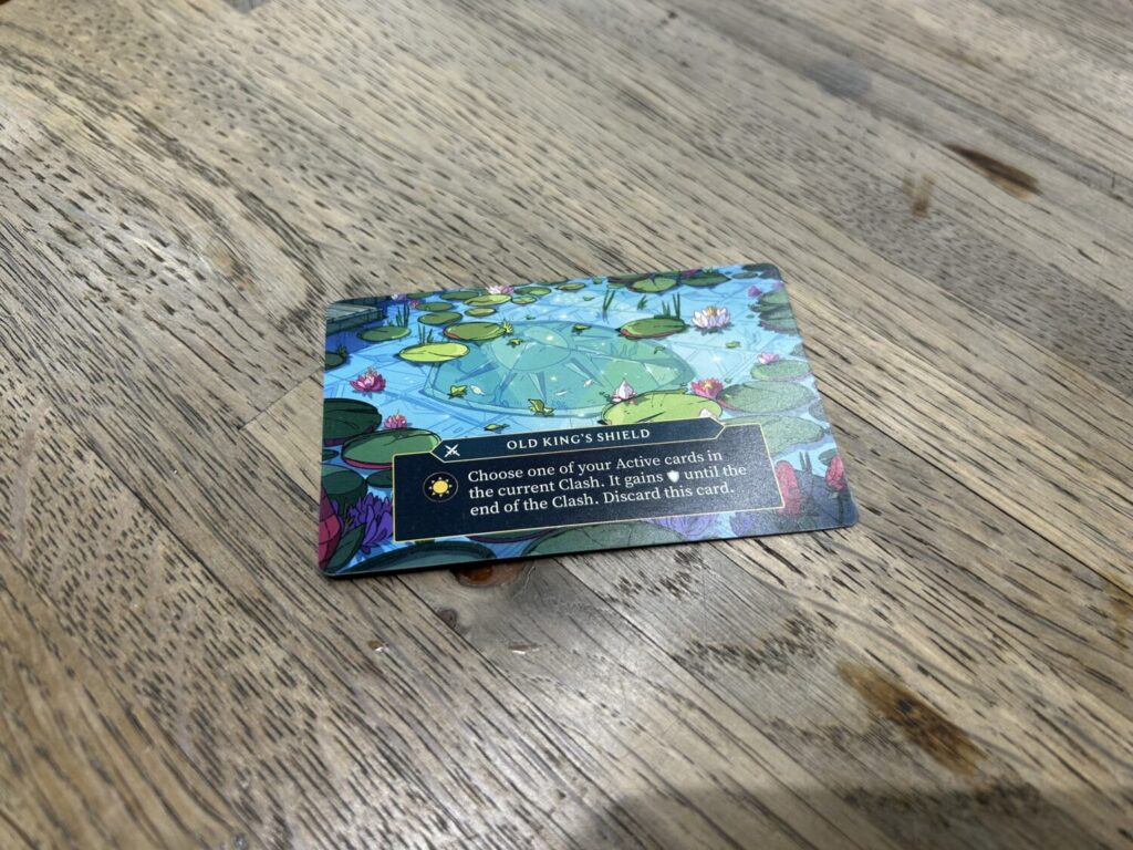 One of the many ability cards that can be found in the game, this one shows a gorgeous illustration of a metal shield sitting at the bottom of a pond, lily pads dotting the surface of the water.