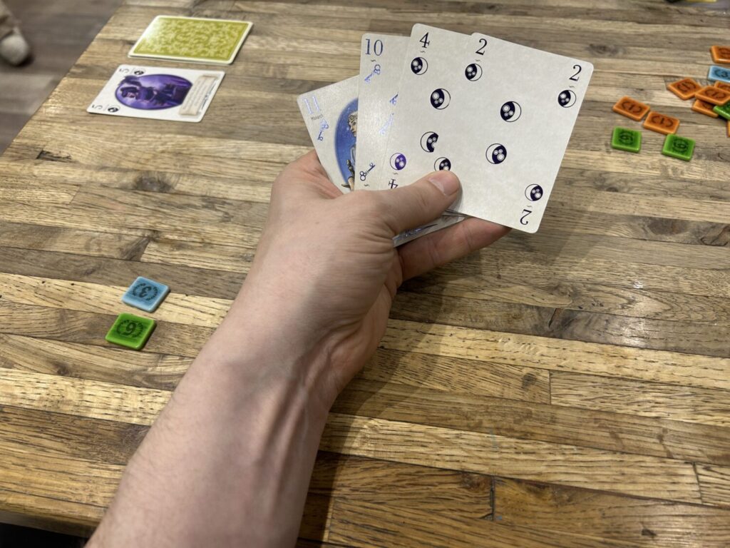 A hand holds several cards above a wooden table. Bakelite scoring tokens sit off to the side.